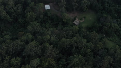 Flying-over-a-thickly-wooded-forest-looking-down-on-a-small-property-tucked-in-the-jungle