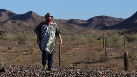 In-the-Sonoran-desert,-a-desperado-man-wearing-a-poncho,-red-bandanna-and-a-cowboy-hat-walks-through-a-rugged-landscape-filled-with-rocks-and-saguaro-cacti