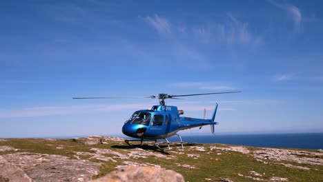Private-helicopter-perched-on-mountain-cliff-summit-overlooking-blue-ocean-landscape-with-propeller-spin-slow-left