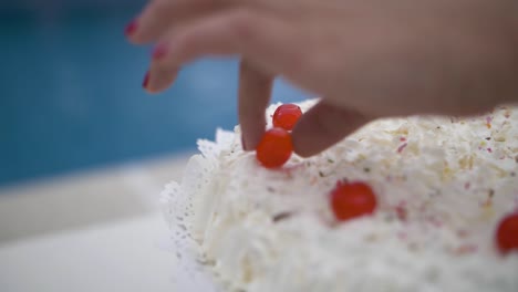 Hand-finishing-sweet-cream-cake-with-red-candy-cherries-on-celebration-event-outdoors-with-swimming-pool-in-the-background