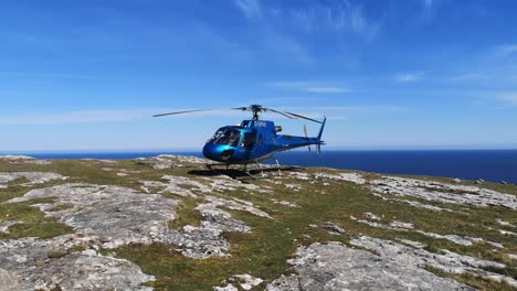Private-helicopter-perched-on-mountain-cliff-summit-overlooking-blue-ocean-landscape-with-starting-propeller-spin
