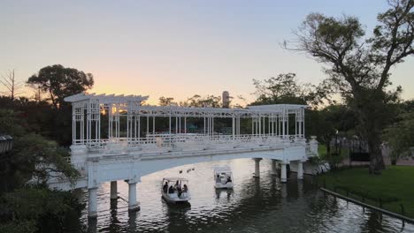 Luftschwenk-Links-Von-Booten,-Die-Unter-Der-Weißen-Brücke-In-Rosedal-Gardens,-Palermo-Viertel-Bei-Sonnenuntergang,-Buenos-Aires-Fahren