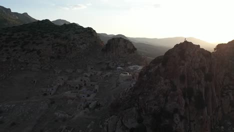 Aerial-shot-moving-to-the-right-and-back-over-the-small-ancient-village-in-ruins,-next-to-a-steep-rocky-mountain-in-the-beautiful-and-warm-sunset-in-Tunisia