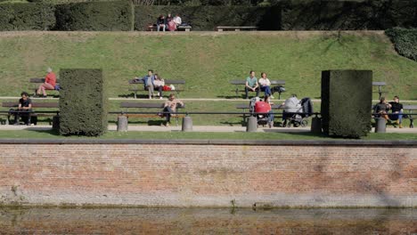 People-Sitting-And-Relaxing-On-Bench-Under-Warm-Sunlight-In-Front-Of-Small-Pond-In-La-Cambre-Abbey-In-Ixelles,-Brussels,-Belgium