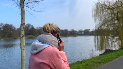 Pretty-young-Caucasian-woman-in-pink-coat-calling-with-smartphone-during-walk-outdoors-at-lake