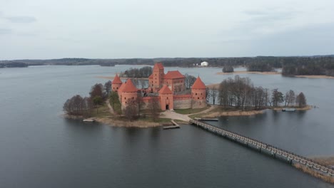 Luftaufnahme:-Burg-Der-Insel-Trakai,-Umgeben-Von-Einem-See-An-Einem-Trüben-Tag-Mit-Holzbrücke,-Die-Die-Nächste-Insel-Verbindet