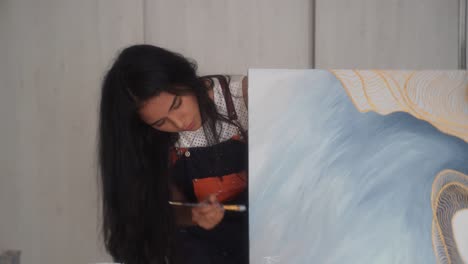 Hispanic-Female-Painter-With-Apron-Brushing-Paint-On-Side-Of-Canvas