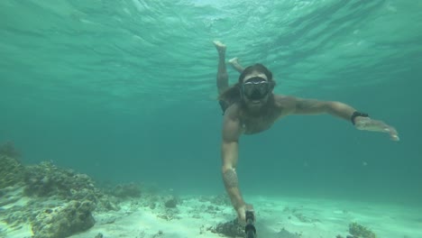 A-young,-fit-and-strong-man-with-long-hair-and-beard-is-swimming-with-snorkeling-goggles-dives-down-from-the-surface-to-the-bottom-of-the-ocean-next-to-the-sand,-rocks-and-reef-and-swims-up-again