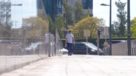 Handheld-shot-of-man-walking-on-sidewalk-away-from-camera-next-to-reflecting-wall-on-sunny-warm-day-in-Brussels,-Belgium