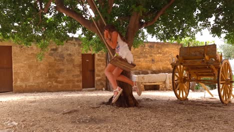 Back-view-of-adorable-girl-on-swing-tied-to-tree-with-Sicilian-cart-or-carretto-siciliano-in-Sicily,-Italy