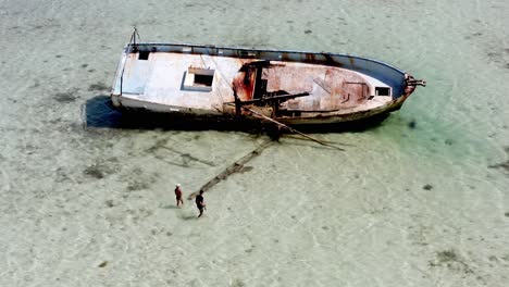 Aerial-View-Of-Two-People-Looking-At-Rusting-Shipwreck-Lies-On-Shallow-Water-Of-Beach