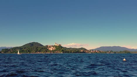 Pequeño-Velero-Navegando-En-El-Lago-Maggiore-Aguas-Agitadas-Con-Montañas-Y-Castillo-De-Angrya-En-El-Fondo,-Italia