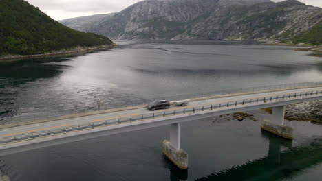 Toma-Aérea-De-Un-Puente-Mientras-Un-Automóvil-Pasa-Por-Encima-Del-Lago-Y-La-Montaña-Como-Telón-De-Fondo,-Noruega