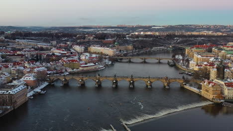 Aerial-view-of-Karluv-Most-and-Prague-cityscape-on-a-winter-evening,-snow-capped-buildings-and-Vltava-river,-Czech-Republic