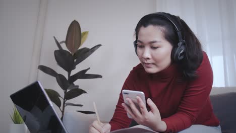 Asiatische-Frau-Auf-Rotem-Hemd,-Die-Ein-Tablet-Mit-Kopfhörer-Für-Ein-Online-Meeting-Zu-Hause-Nutzt