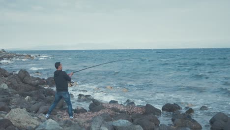 Man-in-difficulty-reeling-in-fishing-rod-along-windy-rocky-shoreline-WIDE-SHOT