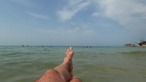 First-person-view-of-male-tourist-legs-relaxing-and-floating-on-inflatable-mattress-with-trabucchi-or-trabocchi-and-people-bathing-in-background