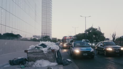 Recent-development-of-Paseo-del-Bajo-complex-with-construction-dumpster-left-on-the-side-of-Eduardo-Madero-Avenue-unattended,-creating-roadside-hazard-during-peak-hours-with-busy-traffic-at-dusk