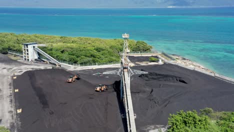 Aerial-orbit-shot-of-working-digger-transporting-coal-at-coal-mine-quarry-in-front-of-paradise-ocean-in-background---Dominican-Republic,Barahona