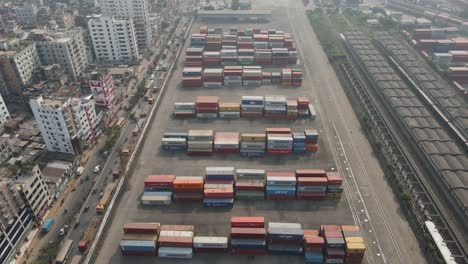 Aerial:-shipping-container-arranged-in-rows-beside-city-highway-and-rail-way-station---drone-flying-top-shot