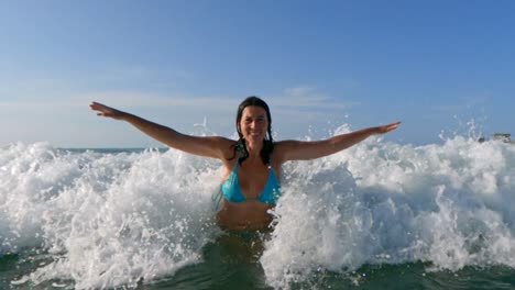 Beautiful-smiling-active-woman-with-blue-bikini-and-arms-wide-open-waiting-big-sea-waves-splashing-on-her