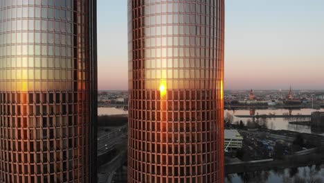Sideway-aerial-drone-view-of-two-high-rise-glass-office-towers-reflecting-golden-sunset-on-windows-with-Riga,-Latvia-cityscape-in-the-background-with-clear-violet-sky