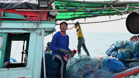 Un-Pescador-Con-Un-Cigarrillo-En-La-Boca-Hablando-Y-Gesticulando-Mientras-Repara-Una-Red,-Muelle-De-Pesca-De-Pattaya,-Chonburi,-Tailandia