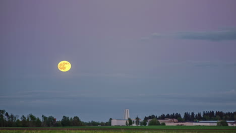 Time-lapse-shot-of-rising-full-moon-on-blue-sky-in-dusk-over-industrial-factory-in-rural-forest-area
