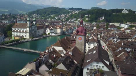 Aerial-orbit-of-Lucerne-Old-Town-picturesque-rooftops,-Jesuit-Church,-Reuss-River-and-bridges,-surrounded-by-hills-and-forest-at-daytime,-Switzerland