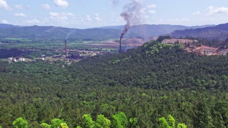 Aerial-forward-over-verdant-landscape-with-smoking-chimneys-in-nickel-mine,-Loma-Miranda