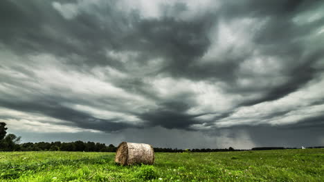 Time-lapse-shot-of-dark-grey-clouds-flying-at-sky-over-agricultural-field-with-hay-bale