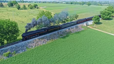 An-Aerial-View-of-a-Steam-Engine-Puffing-Smoke-and-Steam-with-Passenger-Coaches-Traveling-on-a-Single-Track-Going-over-a-Small-Bridge-Thru-Trees-and-Farmland-Countryside-on-a-Spring-Day