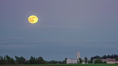 Timelapse-video-of-moon-rising-over-a-blue-sky-in-a-rural-landscape-with-view-of-a-white-building-in-a-distance
