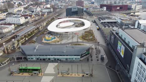 Sandnes-district-court-in-background---Bus-terminal-with-green-bus-and-glass-roof-building-in-foreground---Modern-design-city-square-with-playground-and-ice-skating-arena-in-middle---Aerial-Norway