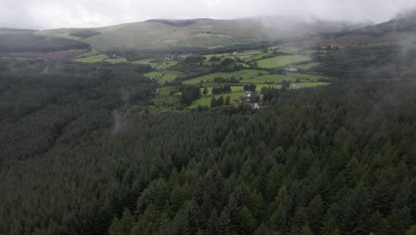 Drone-shot-of-a-green-mountain-valley-in-Ireland-with-fields,-a-forest-and-low-lying-clouds