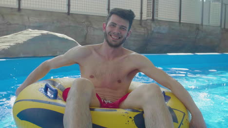 Young-happy-caucasian-male-sitting-on-inflatable-lifebuoy-ring-rowing-in-swimming-pool-water-slow-motion-portrait-of-funny-summer-holiday