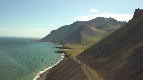 Aerial-orbit-of-hillside-road-near-turquoise-sea,-hills-in-background-in-in-Djúpivogur-small-town-near-Iceland-fiords-at-daytime