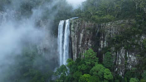 Unique-drone-view-through-suspended-mist-revealing-a-majestic-waterfall-spilling-down-a-lush-rainforest-mountain-scenery