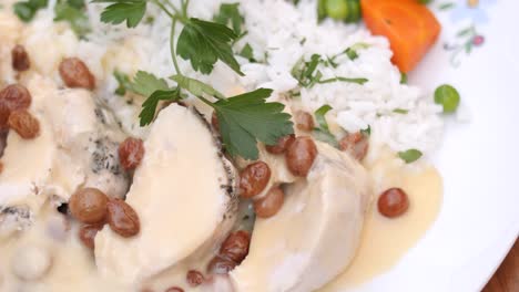Close-up-view-of-rice-with-white-sauce-and-raisins-along-with-boiled-meat-with-carrots-and-peas-and-cilantro-on-the-top-in-a-white-plate-on-a-wooden-table