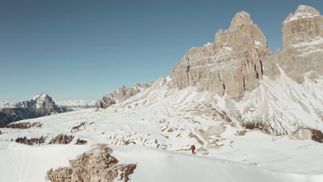 Circling-profile-drone-shot-of-trekker-walking-along-a-steep-snowy-ridge-dolomites-tre-cime-in-the-background