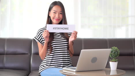 Mujer-Asiática-Feliz-Trabajando-En-Línea-Desde-Casa-Mostrando-El-Mensaje-Hashtag-De-Quedarse-En-Casa,-Cámara-Lenta