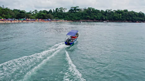 Aerial-tracking-shot-of-small-speedboat-sailing-on-Ubatuba-beach,-Brazil,-heading-towards-crowded-beach