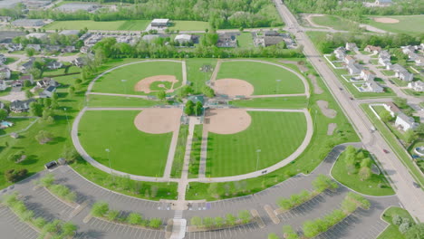 Daytime-Aerial-Shot-Above-Baseball-Diamonds-and-Green-Fields,-Houses,-Road-and-Parking-Lot-Below