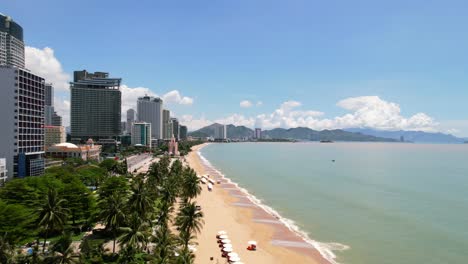 low-altitude-aerial-view-of-a-white-sand-tropical-beach-town-in-asia-with-white-beach-umbrellas-setup-for-tourists-and-tall-hotels-overlooking-the-turquoise-ocean-on-a-sunny-day-in-Nha-Trang-Vietnam