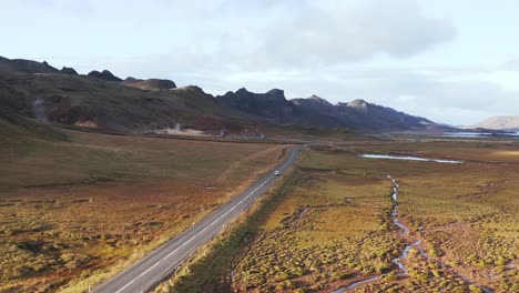 Golden-sunshine-illuminating-wild-tundra-valley-in-Iceland-with-car-traveling-on-road,-aerial