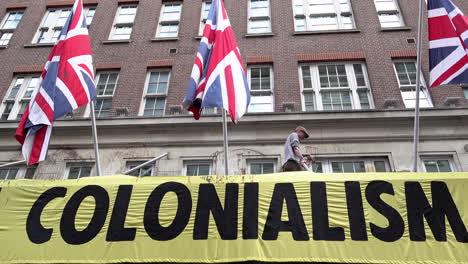 Extinction-Rebellion-climate-change-rooftop-protestors-stand-over-a-yellow-banner-that-reads,-“Colonialism”-and-between-three-British-flags-that-have-been-covered-in-fake-oil