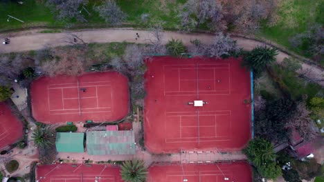 Static-aerial-overhead-view-of-red-clay-tennis-courts-with-people-playing-a-match,-O'Higgins-Park,-Santiago,-Chile