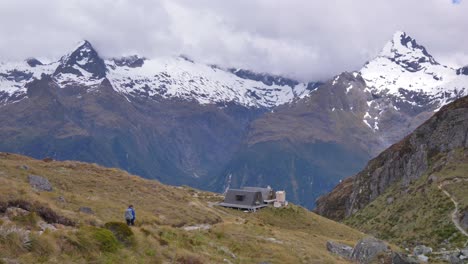 Pan,-hiker-walks-towards-remote-alpine-shelter,-distant-snow-capped-mountains,-Routeburn-Track-New-Zealand