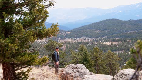 A-solo-male-hiker-walks-out-to-a-viewpoint-and-stops-to-admire-the-view-as-wind-moves-the-trees-around-him