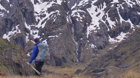 Static,-hiker-walks-past-distant-waterfall-in-snow-capped-landscape,-Routeburn-Track-New-Zealand